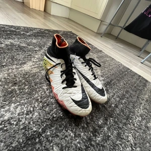 Nike fotbollsskor  - Snygga fotbollsskor från Nike med högt skaft och snörning. Ovandelen är vit med svarta detaljer och stora Nike-loggor. Sulan har ett färgstarkt mönster i gult, orange och rosa med svarta inslag. Perfekta för dig som vill sticka ut på planen. Priset är inte hugget i sten