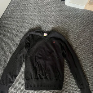 Svart v-ringad tröja J.Lindeberg M - Svart v-ringad tröja från J.Lindeberg med diskret röd logga på bröstet. Långärmad modell i mjuk ullblandning, perfekt för lager-på-lager. Klassisk och stilren med ribbade muddar och nederkant.