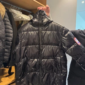 Svart pufferjacka från Canada Goose - Säljer en svart pufferjacka från Canada Goose med klassisk logga på ärmen. Jackan har hög krage, huva och dragkedja framtill. Den är vadderad och har en glansig finish som ger en cool look. Perfekt för kalla dagar när du vill hålla stilen.