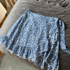 Blå blommig omlottkjol med volang - Supersöt blå kort omlottkjol med vitt blommönster och volangkant. Kjolen har knytband i sidan. I storlek L