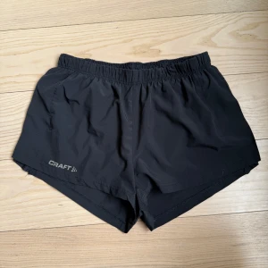 Svarta träningsshorts från Craft - Svarta träningsshorts från Craft i lätt och snabbtorkande syntetmaterial. De har elastisk midja, innerficka och diskreta reflexdetaljer på sidorna. Perfekta för löpning eller gym och har en klassisk sportig look.
