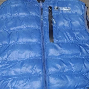 Blå dunväst från Geographical Norway - Snygg blå dunväst från Geographical Norway med quiltad design och svarta detaljer. Västen har dragkedja framtill, en bröstficka med dragkedja och kontrastfärgad insida i svart. Perfekt för lager-på-lager och chill stil.
