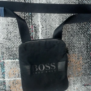 Svart axelväska från Hugo Boss - Snygg svart axelväska från Hugo Boss med justerbart band och dragkedja. Väskan har stilren design med BOSS-logga framtill och på dragkedjan. Perfekt storlek för mobil, plånbok och andra småsaker. Tillverkad i slitstarkt syntetmaterial.