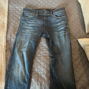 Blå jeansbyxor från jack and jones, stl 31/32 - Säljer ett par snygga blå jeans  i storlek 31/32. Jeansen har klassisk femficksdesign, tydliga slitningar framtill och bakfickor med diskreta sömmar. Mörk tvätt med ljusare partier för en cool, använd look. Perfekta till sneakers eller boots.