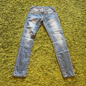 True Religion ljusblå jeans, stl 27 - Säljer ett par klassiska ljusblå jeans från True Religion med snygga kontrastsömmar och ikoniska broderade bakfickor. Modellen har raka ben och låg midja. Jeansen är i bomull och har detaljerade knappar med logga. Perfekta för dig som gillar street och premium denim.