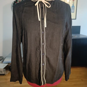Svart blus med knyt och vita detaljer - Svart långärmad blus med vita kantband längs knäppningen och kragen. Söt knytning vid halsen och svarta knappar framtill. Skjortan har en klassisk passform och är tillverkad i ett mjukt bomullstyg. Perfekt för dig som gillar stilrena detaljer.