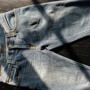 Blå jeans från Nudie Jeans - Säljer ett par klassiska blå jeans från Nudie Jeans med snygga kontrastsömmar och ikonisk broderad bakficka. Jeansen har normal passform, fem fickor och är tillverkade i slitstarkt denim. Perfekta för dig som gillar en tidlös och avslappnad stil.
