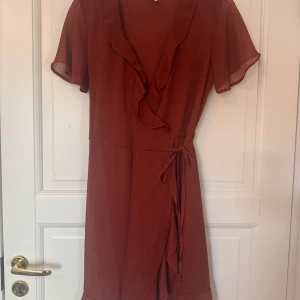 Omlottklänning med volang - Supersöt kortklänning med omlottmodell och knyt i midjan. Klänningen har volangdetaljer längs urringningen och nederkanten samt korta, luftiga ärmar. Perfekt för dig som gillar en feminin och trendig stil.