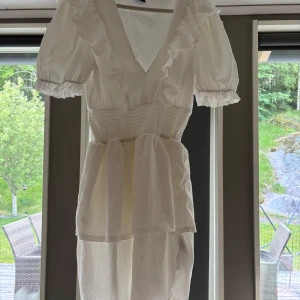 Vit volangklänning med v-ringning - Superfin vit klänning med v-ringning och volangdetaljer längs halsen. Puffiga korta ärmar med resår och smock i midjan för en snygg passform. Lätt och luftig, perfekt för varma dagar. Materialet är strukturerat och känns som bomull. OBS: lite sönder vid lappen med går att laga 