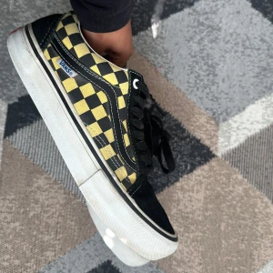 Vans Old Skool gul-svart checkerboard - Snygga Vans Old Skool sneakers med klassiskt checkerboard-mönster i gult och svart. Skorna har svart mocka framtill och på hälen, vita sidoränder och svart snörning. Tjock vit sula och ikonisk Vans-logga på sidan. Perfekta för en cool streetstil.