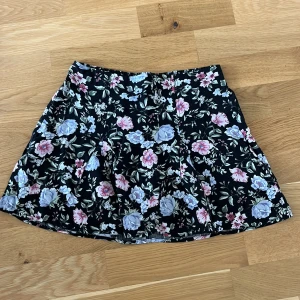 Blommig kortkjol från H&M Divided - Supersöt svart kortkjol från H&M Divided med blommigt mönster i lila, rosa och vitt. Kjolen har en lätt utställd form och resår i midjan för extra bekvämlighet. Perfekt för dig som vill ha en feminin och trendig look.
