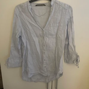 Ljusblå linneskjorta från Zara Basic - Säljer en ljusblå skjorta i linne från Zara Basic Collection. Skjortan har v-ringning, knappar framtill, bröstficka och trekvartsärmar med snörning vid ärmsluten. 