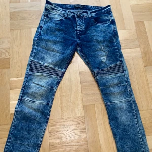 Philipp Plein blå jeans - Säljer ett par blå jeans från Philipp Plein med cool tvättad effekt och ribbade detaljer över knäna. Jeansen har fem fickor, raka ben och metallpatch bak. Perfekt för dig som vill sticka ut med en edgy streetstil.