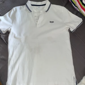 Vit pikétröja från Hugo Boss - Vit pikétröja från Hugo Boss med klassisk krage och knappar framtill. Tröjan har korta ärmar och mörkblå detaljer på krage och ärmslut. Broderad Boss-logga på bröstet. Perfekt för en clean och stilren look.