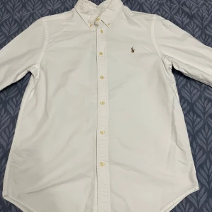 Vit skjorta från Ralph Lauren - Klassisk vit skjorta från Ralph Lauren med broderad logga på bröstet. Skjortan har lång ärm, knappar framtill och button-down krage. Snygg och tidlös modell i bomull som passar perfekt till jeans eller chinos. knappast använd och har inga fläckar eller så. nya orginal priset: 1.795kr. skriv privat om frågor!!