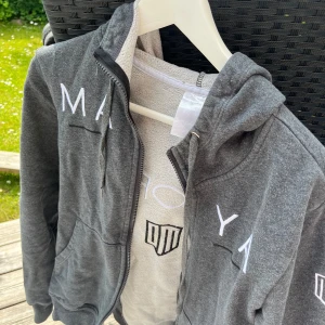 Grå hoodie med dragkedja från Delorez - Mörkgrå hoodie från Delorez med vit broderad logga och text både fram och bak. Tröjan har huva, snörning och dragkedja framtill samt två fickor. Perfekt för chill dagar eller när du vill ha en sportig look.