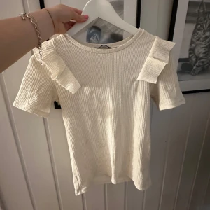 Ribbad beige topp med volangdetaljer - Superfin ribbad topp i beige med korta ärmar och volangdetaljer på axlarna. Perfekt för dig som gillar en söt och stilren look. Mjuk och skön, passar till både jeans och kjol.(har en liten fläck)