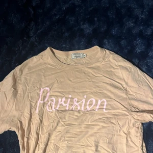 Beige Parisien t-shirt Maison Kitsuné - Snygg beige t-shirt från Maison Kitsuné med rosa texten 'Parisien' framtill. Klassisk rund halsringning och korta ärmar. Tillverkad i mjuk bomull som känns skön mot huden. Perfekt för dig som gillar stilrena och trendiga plagg med en fransk vibe.
