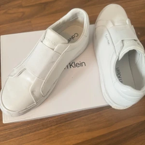 Vita sneakers från Calvin Klein - Stilrena vita sneakers från Calvin Klein med elastiskt band över vristen istället för klassisk snörning. Skorna har en minimalistisk design med rund tå och är tillverkade i skinn. Perfekta för en clean och modern look.