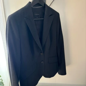 Svart kavaj från Stockh LM, stl 42 - Klassisk svart kavaj från Stockh LM i storlek 42. Insvängd passform med två knappar framtill, spetsiga slag och fickor med lock. Perfekt för en stilren look. Tunt fodrad insida för extra komfort.