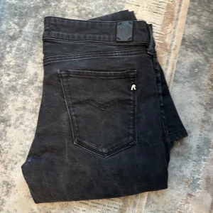 Svarta jeansbyxor från Replay - Säljer ett par svarta Replay jeansbyxor i modell Anbass, storlek W30 L30. Byxorna har klassisk femficksdesign, raka ben och diskret Replay-logga på bakfickan. Perfekta för en stilren och enkel look.