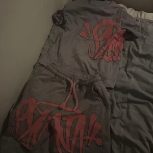 Syna world set - Grå t-shirt från syna world med stor rosa graffiti-inspirerad print på både fram- och baksidan. T-shirten är kortärmad och har en avslappnad passform. Materialet är mjuk bomull, perfekt för en chill streetwear-look.