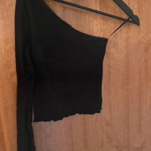 Svart oneshoulder croppad topp - Snygg svart croppad topp med oneshoulder-design och lång ärm. Toppen har en stilren look och lätt vågig kant nedtill. Perfekt för dig som vill sticka ut med en cool och trendig stil.
