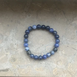 Blått pärlarmband med stenar - Snyggt armband med runda pärlor i olika blå nyanser, vissa med vita inslag. Pärlorna är trädda på elastisk tråd och en av pärlorna är metallisk och fyrkantig, vilket ger en cool kontrast. Perfekt accessoar för att lyfta din stil.