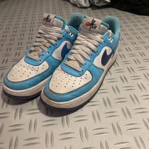 Nike Air Force 1 blå och vita sneakers - Nike Air Force 1 sneakers i en fräsch kombination av ljusblått och vitt med mörkblå swoosh. Klassisk låg modell med snörning, perforerad tå och platt sula. Ovandel i skinn och textilfoder för extra komfort. Perfekta för dig som gillar retrovibbar och streetstyle.