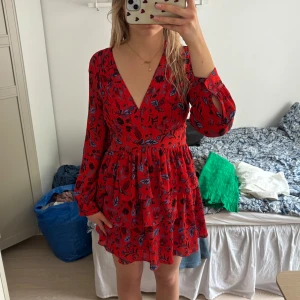 Röd blommig kortklänning - Supersöt röd kortklänning med blommigt mönster i blått och svart. Klänningen har v-ringning, långa ärmar med slits och volangig kjol. Perfekt för dig som gillar färg och vill sticka ut lite extra.