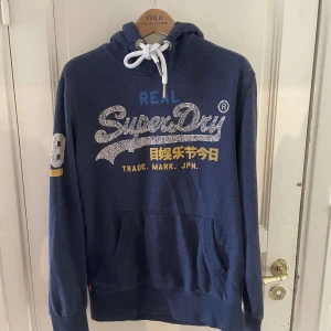 Blå Superdry hoodie med tryck - Mörkblå hoodie från Superdry med stort glittrigt logotryck och japanska tecken på bröstet. Tröjan har huva med vit snörning, känguruficka och tryckt siffra på ärmen. Perfekt för en chill och sportig look.