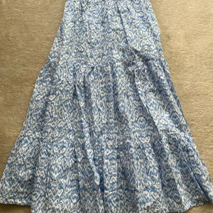 Blåvit långkjol från Lindex - Långkjol från Lindex i storlek 146 med blått och vitt abstrakt mönster. Kjolen har volanger och resår i midjan för en skön passform. Tillverkad i mjuk bomull, perfekt för varma dagar.