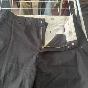Svarta Dickies chinos straight fit - Svarta chinos från Dickies med raka ben och klassisk passform. Byxorna har bälteshällor, dragkedja och knapp framtill samt två bakfickor med knapp. Materialet är slitstark bomull och de har en clean, tidlös look.