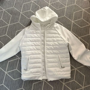 Vit cardigan  - Snygg vit pufferjacka med quiltade partier och dragkedja framtill. Jackan har huva, två fickor med dragkedja och raka ärmar. Perfekt för dig som gillar en clean och stilren look. Materialet känns mjukt och lätt, passar bra till kyligare dagar.