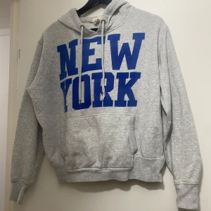 Grå hoodie med blå New York-tryck H&M - Grå hoodie från H&M i storlek S med stort blått 'NEW YORK'-tryck på bröstet. Klassisk känguruficka, snörning i huvan och ribbade muddar. Perfekt för chill dagar eller när du vill ha en sportig streetstyle.