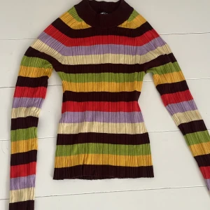 Randig ribbad polotröja Gina Tricot - Färgglad ribbad polotröja från Gina Tricot i storlek XS. Tröjan har breda ränder i vinrött, gult, grönt, lila, rött och beige. Den är långärmad och har en tight passform med hög krage. Perfekt för dig som gillar retrovibbar.