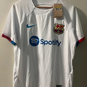 Vit FC Barcelona Nike Dri-FIT tröja - FC Barcelona fotbollströja från Nike i vitt med blå och röda detaljer på ärmsluten. Tröjan har klubbmärke, Nike-logga och Spotify som sponsor framtill samt UNHCR-tryck på ryggen. Tillverkad i Dri-FIT-material som andas och håller dig torr.