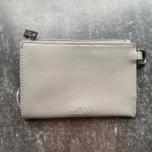 Grå plånbok från BOSS - Stilren grå plånbok från BOSS i slätt skinn med diskret logga framtill. Praktisk dragkedja upptill och metallfäste i sidan för enkel användning. Perfekt för kort och kontanter, minimalistisk design som passar till allt.