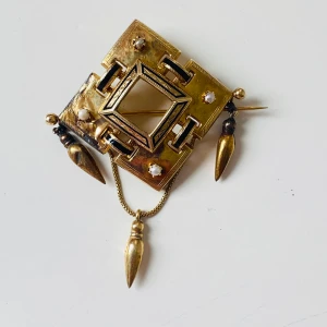 Vintage brosch i guldton med pärlor - Unik fyrkantig brosch i guldtonad metall med geometriskt mönster, svarta detaljer och små vita pärlor. Hängande droppformade hängen och kedja ger extra edge. Perfekt accessoar för dig som gillar statement-smycken med retrovibbar.