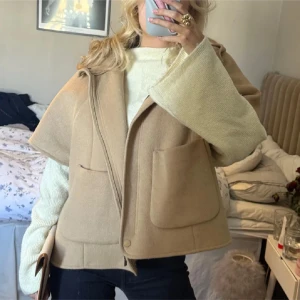 Beige capejacka med huva från Zara - Säljer en beige capejacka från Zara med huva och stora fickor framtill. Jackan har korta ärmar, knappar vid sidorna och är tillverkad i ett mjukt ullblandat material. Perfekt för lager-på-lager och en snygg detalj till din outfit. Storlek 164, motsvarar en S!😊två första bilderna är inte mina
