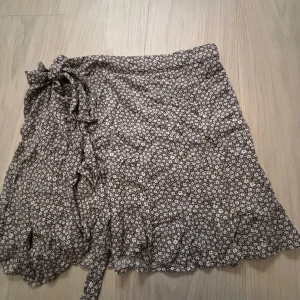 Blommig kjol från BikBok - Supersöt brun kort omlottkjol från BikBok med små vita och gula blommor. Kjolen har volangkant och knyt i sidan. Går att justera hur tight du vill ha den!
