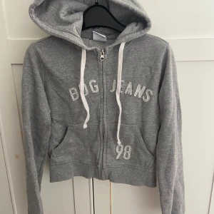 Grå hoodie från BDG Urban Outfitters - Grå hoodie med dragkedja från BDG Urban Outfitters. Broderad text 'BDG JEANS' och siffran 98 framtill, vita snören i huvan och två fickor. Mjuk bomullsblandning och ribbade muddar. Perfekt för en chill och avslappnad stil. Litet hål under luvan men märks inte när det är på.