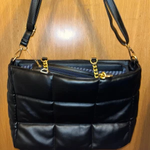 Axelväska - Snygg svart axelväska i skinnimitation med guldfärgade kedjedetaljer och justerbar axelrem. Väskan har flera fack med dragkedja och ett randigt innerfoder. Perfekt för dig som vill ha en stilren och trendig väska med plats för allt du behöver 🤍.