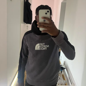 Grå hoodie från The North Face - Säljer en grå hoodie från The North Face med svart huva och svart detalj längs ena ärmen. Stor vit logga på bröstet. Hoodien har en klassisk passform och är tillverkad i mjukt material, perfekt för kyliga dagar.