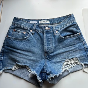 Blå jeansshorts från Mango  - Snygga blå jeansshorts från MNG med hög midja och råa, slitna kanter. Shortsen har klassisk femficksdesign och coola slitningar framtill för en avslappnad vibe. Perfekta till sommaren och lätta att matcha med allt. De är en storlek 34, och jag som vanligtvis har 36 får på mig dom, men de sitter lite tajt. Midjan är medelhög