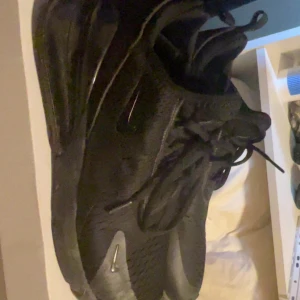 nike 270 - Säljer ett par helsvarta Nike Air Max sneakers med chunky sula och synlig Air Max-dämpning. Skorna har mesh- och syntetmaterial, snörning och diskreta loggor på sidan och hälen. Perfekta för dig som gillar sportig och clean stil.