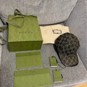 Gucci keps (ÄKTA), storlek M - Snygg keps från Gucci i mörkbrunt tyg med klassiskt GG-monogram i beige. Kepsen har justerbar rem bak i svart skinn och detaljer i svart runt skärmen. Kommer med originalförpackning och dustbag. Perfekt för dig som vill sticka ut med lyxig streetstyle. Först till kvarn som gäller, kostar runt 5 tusen säljs för 2,6 dock är inte priset skrivet på Sten så vid snabbt köp går att justera lite grann (ÄKTA )