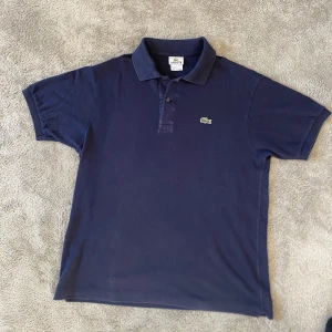 Lacoste piké - Säljer en klassisk mörkblå pikétröja från Lacoste med den ikoniska gröna krokodilloggan på bröstet. Tröjan har korta ärmar, krage och knappar framtill. Perfekt för en stilren och avslappnad look. Modellen är 183 cm och pikén är perfekt för sommaren. Nypriset är cirka 1200 kr!