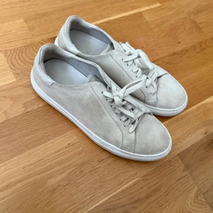 Beige mockasneakers från Axel Arigato - Stilrena sneakers i beige mocka från Axel Arigato. Skorna har rund tå, vita snören och vit platt sula. Perfekta för en clean och minimalistisk look. Klassisk design med diskret logga på sidan.