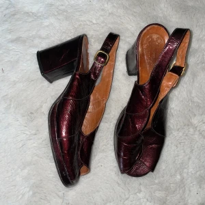 Vinröda retro klackskor i skinn - Snygga vinröda klackskor i skinn (syntetisk ovandel, äkta foderläder) med öppen tå och bred klack. Skorna har justerbart spänne i guld och cool retrodesign med vågiga paneler. Perfekta för dig som gillar statementskor med vintage vibe. Ser ingen storlek, men lite trånga på mig som har 38/39, så skulle säga 36/37. 